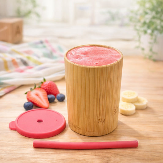 Bamboo cup with pink smoothie, strawberries, blueberries, and bananas on a wooden table.