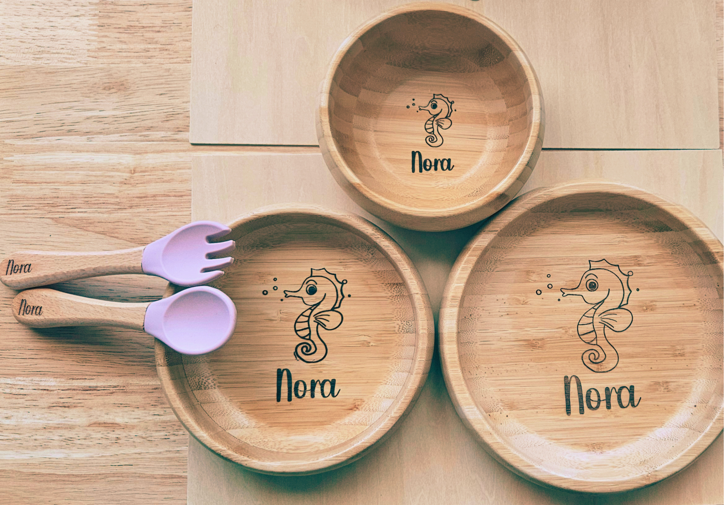 Set of wooden plates and spoons with 'Nora' and seahorse design on a wooden surface.