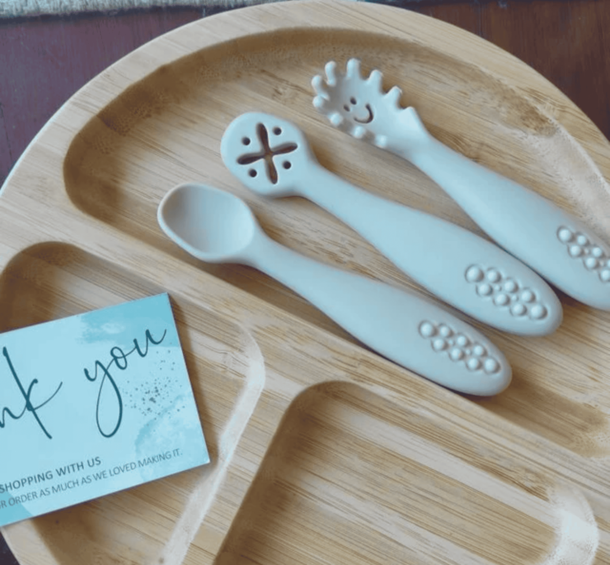 White baby utensils on a wooden tray with a 'thank you' card.
