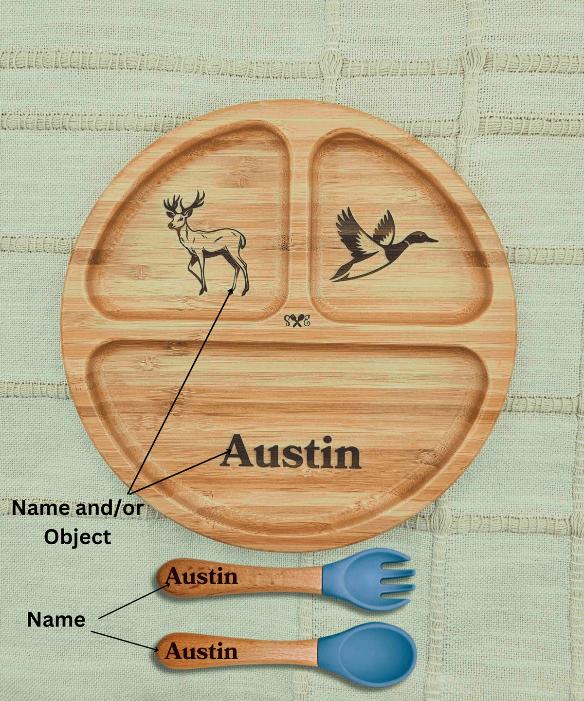 Personalized round wooden divided plate with engraved animal illustrations and the name 'Austin' on a textured light green fabric background, shown with matching wooden utensils and blue spoons below.