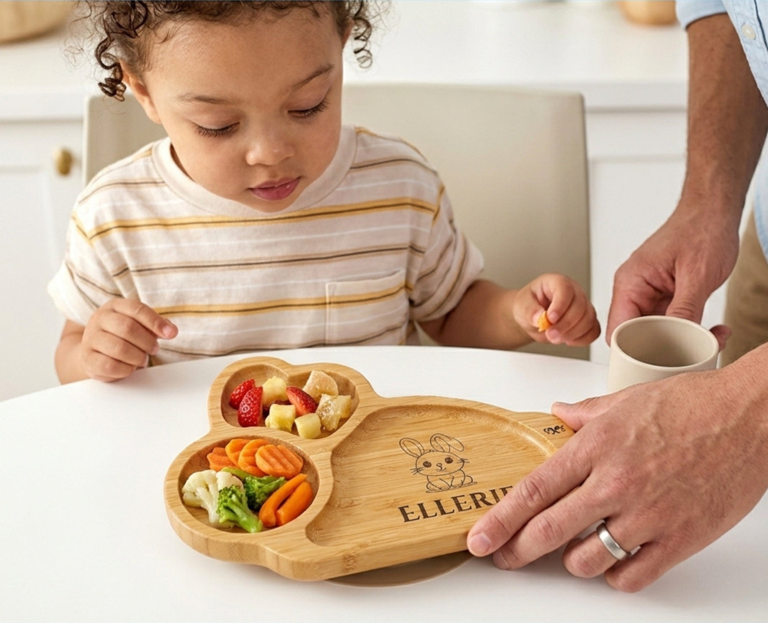 Child sitting at a table with a wooden plate of food, supervised by an adult.